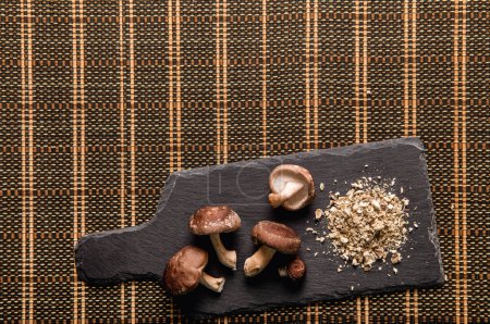 Flache Ansicht von trockenem Pulver aus Shiitake-Pilzen, Lentinula edodes. Lebensmittelzutat auf schwarzem Steinschneidebrett mit frischen Shiitake-Pilzen. Raum für Text.