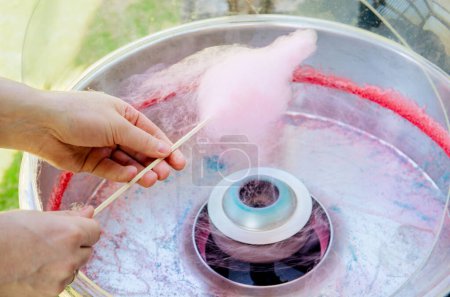 Close up view of person hands spinning pink color cotton candy with machine. Spin around wood stick outdoors in summer.