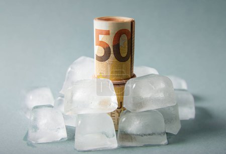 Concept of freezing the funds. Euro money roll inside ice cubes on blue studio background. Copy space.