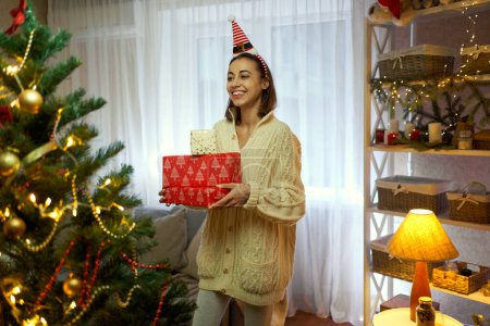 Foto de Chica divertida festiva sosteniendo caja de regalo de Navidad en la sala de estar por sala de estar decorada. Navidad y Año Nuevo - Imagen libre de derechos