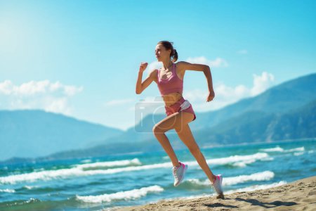 Foto de Focused strong young woman running on sunny beach seaside sprinting fast exercising cardio workout training - Imagen libre de derechos