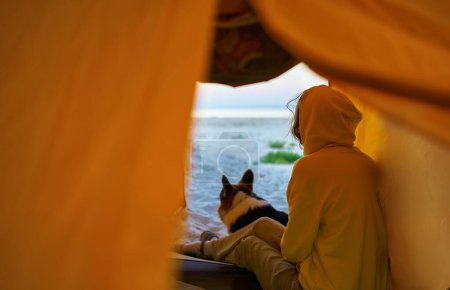 Foto de Mujer viajera y su mascota sentadas dentro de la tienda y admirando la vista de la naturaleza en la playa tranquila. Experimenta la alegría de explorar el mundo con tu peludo amigo - Imagen libre de derechos