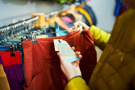 Gros plan de mains féminines tenant une paire de pantalons rouges avec une étiquette de prix, symbolisant le concept de shopping et de consumérisme