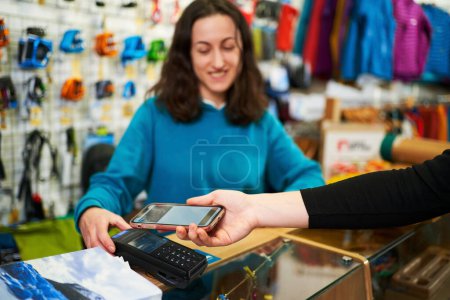 Foto de Cliente haciendo el pago con teléfono inteligente a través de una terminal en la tienda. La vendedora acepta el pago detrás del mostrador. - Imagen libre de derechos