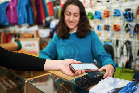 Käufer bezahlen per Smartphone mit leerem Bildschirm über ein Terminal im Geschäft. Die Verkäuferin akzeptiert die Zahlung hinter der Theke