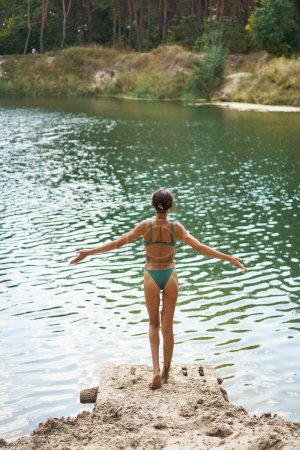 Rückenansicht einer Frau im Bikini mit perfekt gebräuntem Körper genießt den ruhigen See und öffnet die Arme