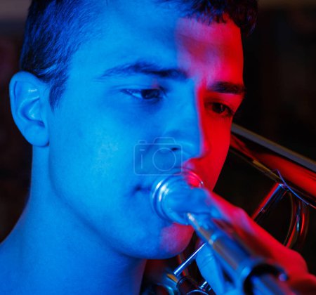 Téléchargez les photos : Jeune homme talentueux jouant trombone avec la lumière bleue et rouge sur son visage - en image libre de droit