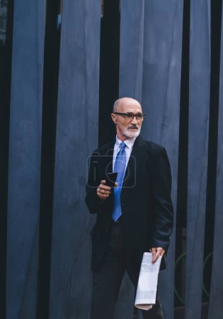 Foto de Hombre maduro de pelo gris confiado y envejecido en ropa formal con periódico y teléfono inteligente saliendo al escenario para realizar un discurso al aire libre - Imagen libre de derechos