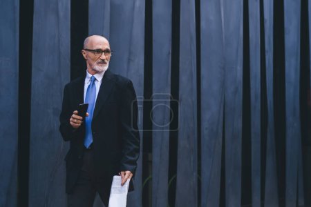 Foto de Hombre maduro de pelo gris confiado y envejecido en ropa formal con periódico y teléfono inteligente saliendo al escenario para realizar un discurso al aire libre - Imagen libre de derechos