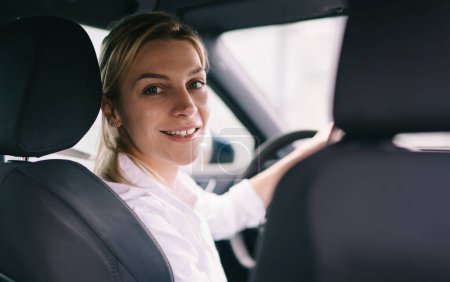 Back view of smiling blond woman in white shirt sitting at steering wheel in car and looking at camera over shoulder