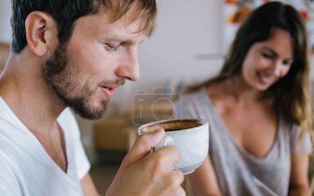 Téléchargez les photos : Accent doux sur beau millénial homme appréciant tasse de café chaud acidulé tandis que la femme floue sur fond écriture sur papier et souriant - en image libre de droit