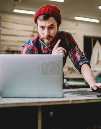 Contemporary bearded male joiner in casual colorful clothing and red hat watching video on laptop while working on project at workshop
