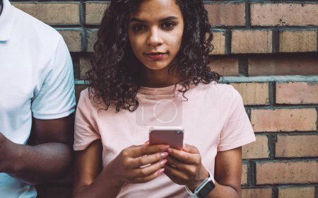 Photo for Half length portrait of curly hipster girl dressed in casual wear holding cellular gadget in hands and looking at camera near promotional background while boyfriend ignoring live communication - Royalty Free Image