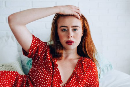 Photo for Afraid attractive woman holding red hair and being sorry for text on smartphone while lying on bed in hotel room - Royalty Free Image
