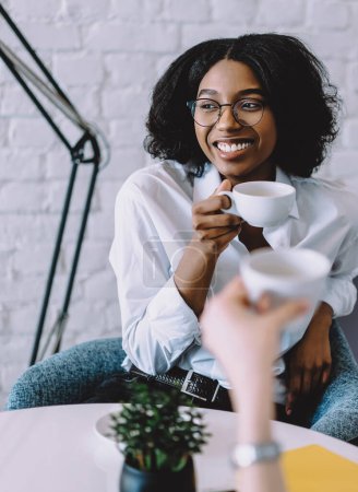 Photo for Cute positive dark skinned young woman holding cup of coffee in hand and communicating with best friend during meeting in modern apartment.Pretty african american hipster girl laughing with colleague - Royalty Free Image