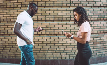 Photo for African American hipster guys chatting on smartphone gadgets in social networks while walking on urban setting, millennial generation using mobile phones for communication and online messaging - Royalty Free Image