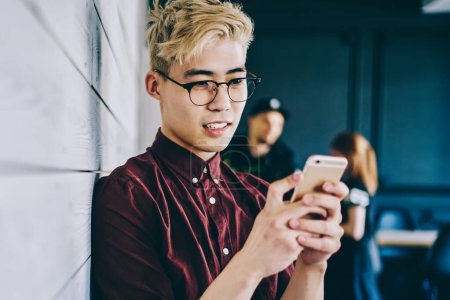 Photo for Chinese hipster blogger in eyeglasses reading incoming notification on smartphone standing near publicity area for your advertisement.Asian young man chatting online on mobile phone - Royalty Free Image