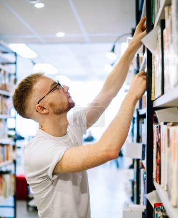 Téléchargez les photos : Vue latérale de l'homme barbu adulte coûteux en chemise blanche et des lunettes de mise au point tout en choisissant le livre sur l'étagère dans la bibliothèque moderne lumière - en image libre de droit