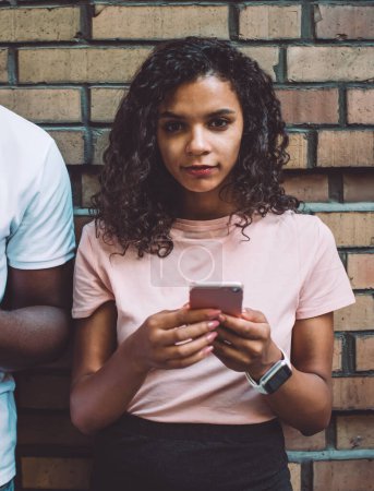 Photo for Half length portrait of curly hipster girl dressed in casual wear holding cellular gadget in hands and looking at camera near promotional background while boyfriend ignoring live communication - Royalty Free Image