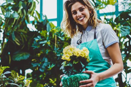 Half length portrait of female florist looking at camera with wide smile on face and enjoying time for caring of flowers , happy botanist holding pot with blooming plant in hands in orangery