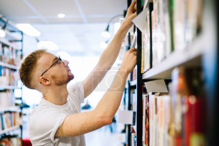 Téléchargez les photos : Vue latérale de l'homme barbu adulte coûteux en chemise blanche et des lunettes de mise au point tout en choisissant le livre sur l'étagère dans la bibliothèque moderne lumière - en image libre de droit
