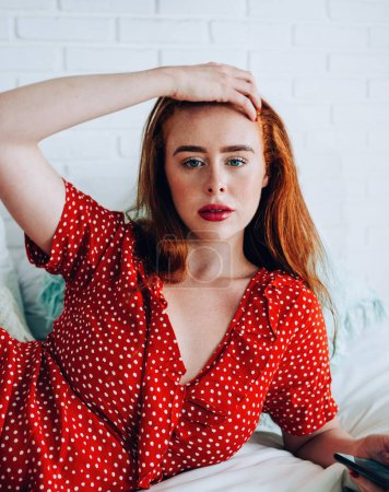 Photo for Afraid attractive woman holding red hair and being sorry for text on smartphone while lying on bed in hotel room - Royalty Free Image