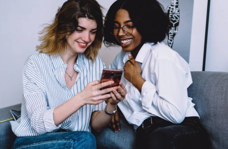 Photo for Cheerful african american blogger with positive hipster girl laughing while viewing common photos on smartphone using home internet.Diverse friends relaxing together on couch and watching funny video - Royalty Free Image