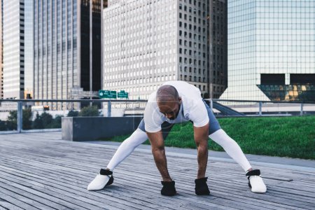 Foto de Hombre afroamericano fuerte atlético en piernas deportivas que estiran las piernas con concentración en la acera de madera cerca de los rascacielos por la mañana mirando hacia atrás. - Imagen libre de derechos