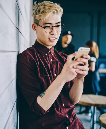 Photo for Chinese hipster blogger in eyeglasses reading incoming notification on smartphone standing near publicity area for your advertisement.Asian young man chatting online on mobile phone - Royalty Free Image