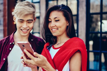 Photo for Positive chinese young man and woman reading funny notification in social networks on modern smartphone standing in urban setting.Cheerful hipster bloggers laughing while watching video on mobile - Royalty Free Image