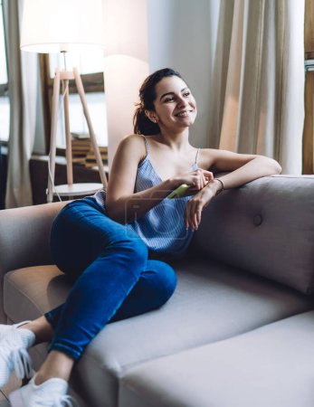 Smiling young lady in tank top and jeans resting on couch and leaning on sofa cushion while playing with bracelet and looking away in apartment