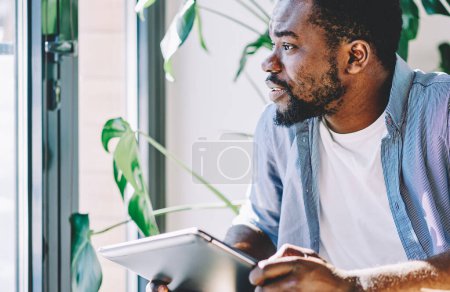 Foto de Afroamericano concentrado freelancer tranquilo mirando por la ventana cuidadosamente mientras sostiene tableta moderna en las manos durante el trabajo a distancia en el espacio de trabajo - Imagen libre de derechos