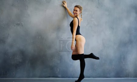 Téléchargez les photos : Vue latérale du contenu mince et attrayant athlète féminine en maillot de bain sport noir et chaussettes de genou penchées avec la main levée sur le mur dans un studio de fitness pendant l'entraînement aérobie en regardant la caméra - en image libre de droit