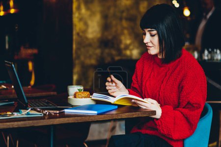 Foto de Encantadora morena joven satisfecha en suéter rojo chequeando horario en notebook mientras trabajaba y sentaba mesa con laptop en cafetería - Imagen libre de derechos