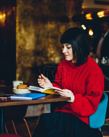 Foto de Encantadora morena joven satisfecha en suéter rojo chequeando horario en notebook mientras trabajaba y sentaba mesa con laptop en cafetería - Imagen libre de derechos