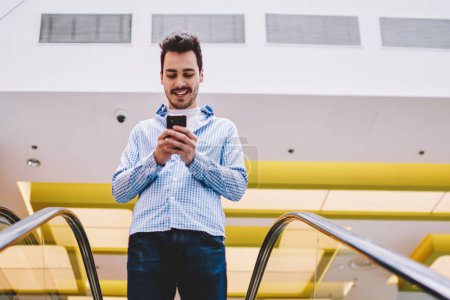 Photo for Positive cheerful hipster guy updating software on cellular phone while standing on escalator and going down for shopping in center, happy man browsing internet on mobile gadget during leisure time - Royalty Free Image