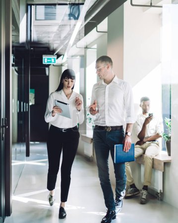 Photo for Enterprising smart multiethnic coworkers in stylish formal wear discussing business while walking on hallway during break in modern office with minimalist interior - Royalty Free Image