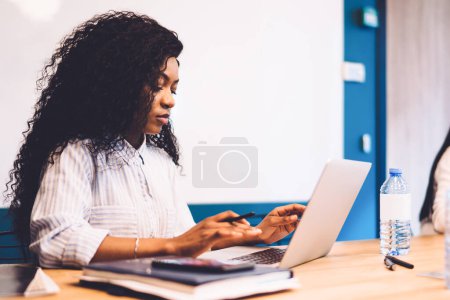 Photo for Focused African American female office worker with long curly hair using portable computer while sitting on wooden table in conference room - Royalty Free Image