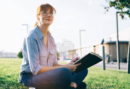 Photo for Wistful red haired female student in casual wear and eyeglasses sitting with legged cross on green grass while working with textbooks on project and looking away - Royalty Free Image
