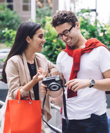 Téléchargez les photos : Contenu souriant couple multiethnique avec sac à provisions reposant sur la rue mégapole tout en regardant appareil photo en plein jour sur fond urbain flou - en image libre de droit