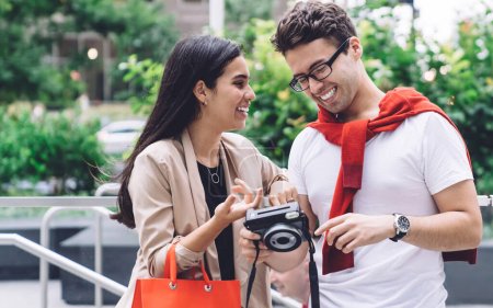 Téléchargez les photos : Contenu souriant couple multiethnique avec sac à provisions reposant sur la rue mégapole tout en regardant appareil photo en plein jour sur fond urbain flou - en image libre de droit
