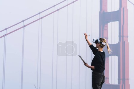 Foto de La joven mujer se extiende de la mano con lápiz digital para un toque interactivo sobre el aire mientras se enfrenta a la construcción de puentes con auriculares de realidad virtual. Hipócrita diseñando espacios arquitectónicos - Imagen libre de derechos
