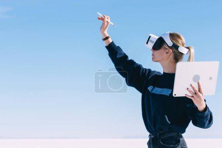 Foto de Las mujeres jóvenes se extienden de la mano con lápiz digital para un toque interactivo sobre el aire, mientras se encuentran en espacios exteriores amplios y limpios con auriculares de realidad virtual. Hipócrita diseñando espacios arquitectónicos - Imagen libre de derechos