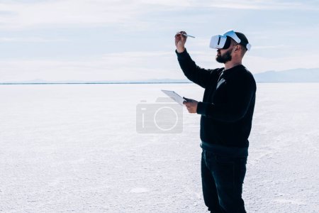 Foto de Guy con lápiz en las manos y tableta digital crea su propia realidad virtual aumentada usando auriculares VR en un amplio paisaje futurista. Arquitecto masculino que construye un proyecto de ciudad en un sitio vacío. - Imagen libre de derechos