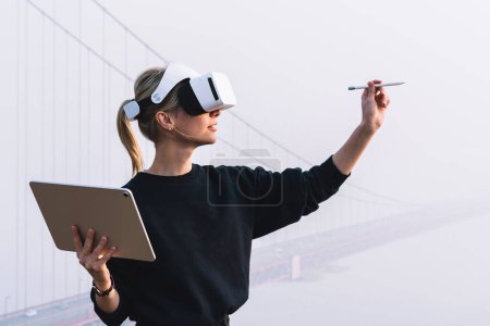 Foto de La joven mujer se extiende de la mano con lápiz digital para un toque interactivo sobre el aire mientras se enfrenta a la construcción de puentes con auriculares de realidad virtual. Hipster chica diseñando espacio de copia de pluma con VR - Imagen libre de derechos