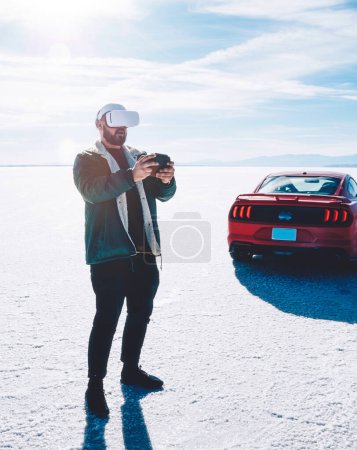 Foto de Tipster guy controlando con joystick simulation world standing near car.Man driving with gamepad playing increased virtual reality dimension game wearing VR headset outdoor in unusual spacious place. - Imagen libre de derechos