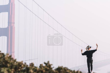 Foto de Mujer en los auriculares de realidad virtual gestación con manos para controlar la simulación de la dimensión futurista 3D de pie al aire libre cerca del puente de la puerta de oro. Hipster niña tocando algo en el mundo de los derechos humanos aumentados - Imagen libre de derechos