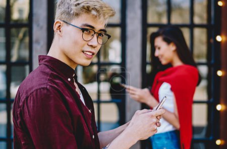 Photo for Handsome chinese hipster guy in eyeglasses looking away while messaging on smartphone and using 4G internet for communication with friend which standing on blurred background with cellular in hand - Royalty Free Image