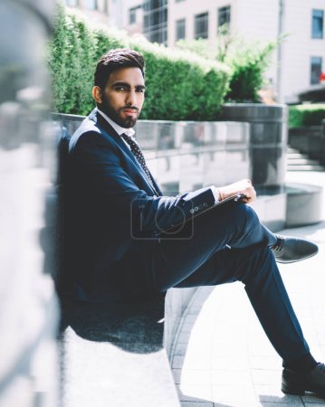 Photo for Portrait of serious spanish corporate director looking at camera while waiting business partner for meeting in financial district, turkish entrepreneur dressed in formal suit sitting outdoors - Royalty Free Image