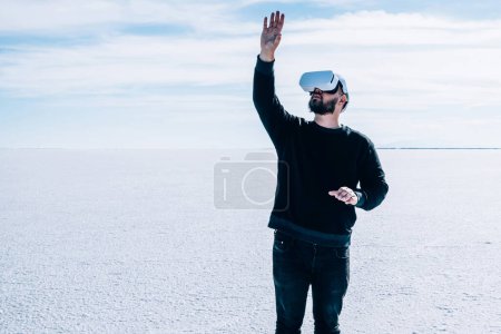 Foto de Hombre en realidad virtual de audífonos gestando con las manos para controlar la simulación de la dimensión futurista 3D mientras se encuentra al aire libre en espacios naturales vacíos. Tipo hipster tocando algo en el mundo de los VR aumentados. - Imagen libre de derechos